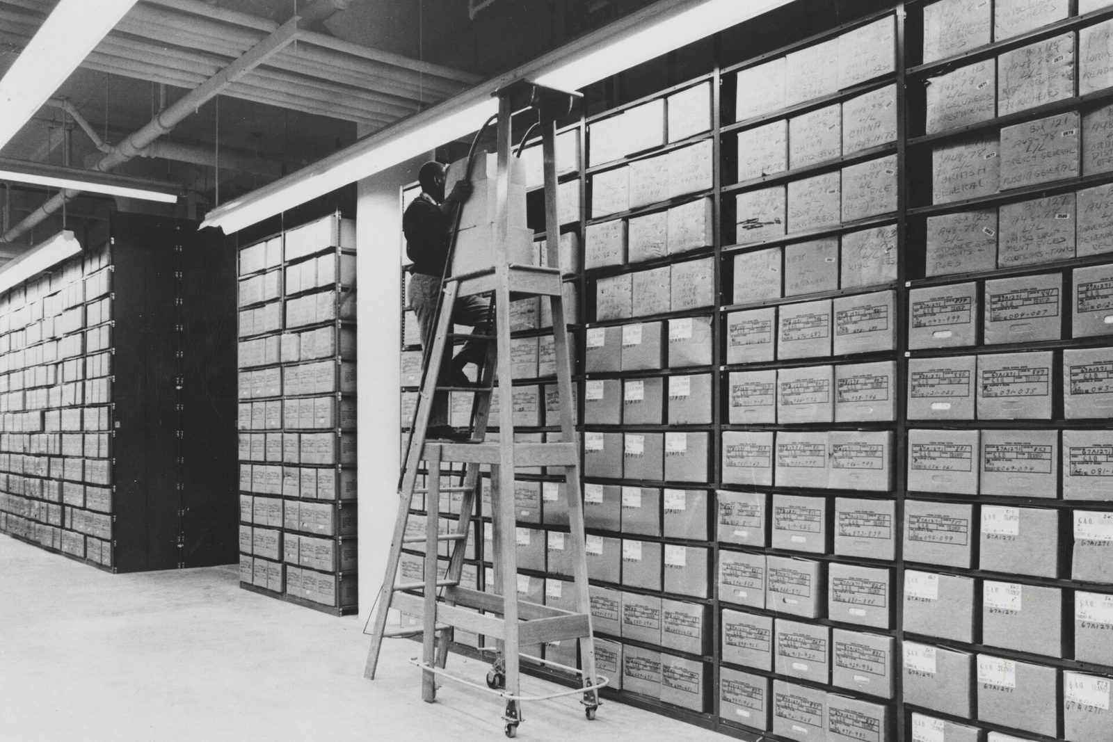 Rows of records boxes on high shelving in a federal records centre, an archivist on a ladder among them